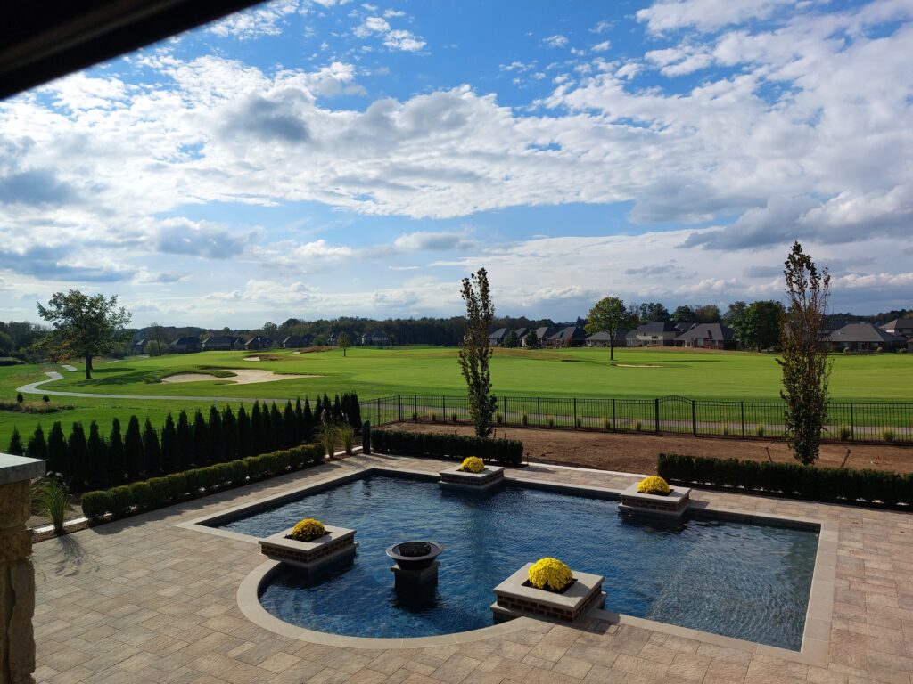 Backyard view featuring a luxurious custom pool with flower planters, surrounded by manicured landscaping and a scenic golf course in the background, emphasizing outdoor living and relaxation.