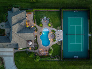 Aerial view of a beautifully designed inground pool surrounded by patio seating and landscaping, adjacent to a tennis court, showcasing the luxurious outdoor space created by Tango Pools.
