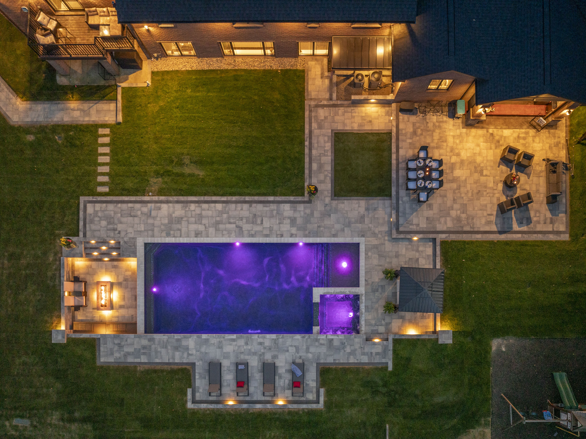 Luxury backyard pool with purple lighting, surrounded by stone patio, outdoor seating, and landscaped lawn, showcasing custom design by Tango Pools.