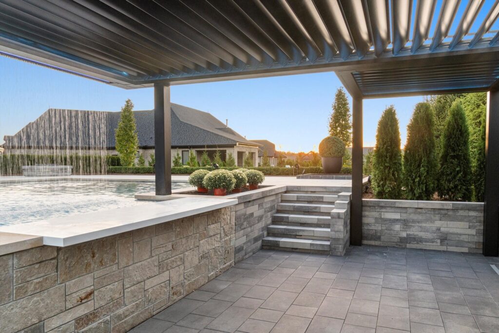 Modern outdoor pool area with a stone wall, steps, lush greenery, and a pergola, showcasing a tranquil oasis designed by Tango Pools.