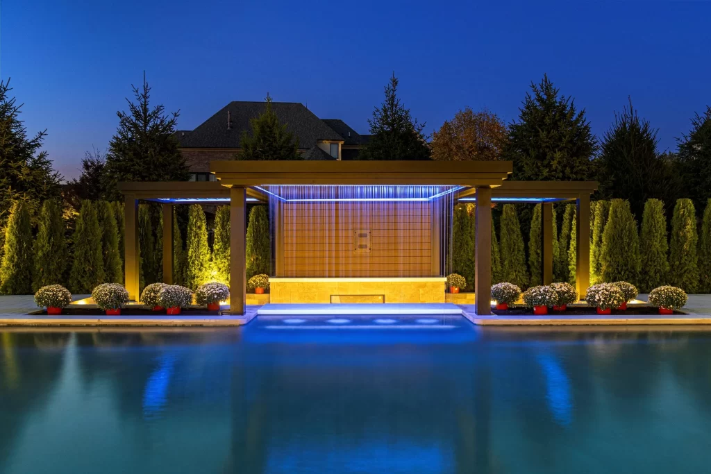 Luxury pool with modern pavilion, illuminated water features, and landscaped greenery, showcasing tile and plaster finishes suitable for Michigan homes.